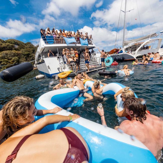 People are enjoying a lively Sydney boat party, swimming and lounging on inflatables in the water, with several boats anchored nearby under a partly cloudy sky.