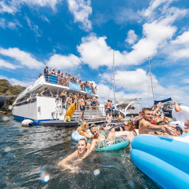 People enjoying a sunny day on yachts and inflatables in the water, surrounded by clear skies and sailboats, making the most of Sydney boat party hire services.