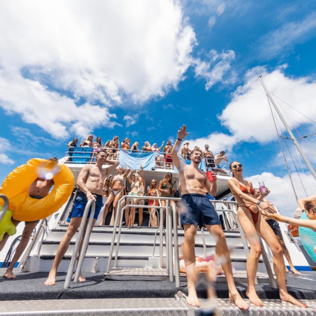 A group of people in swimwear are enjoying a Catamaran party Sydney on a boat with water floats. Some are standing on the deck, while others are on an upper level. It appears to be a sunny day.