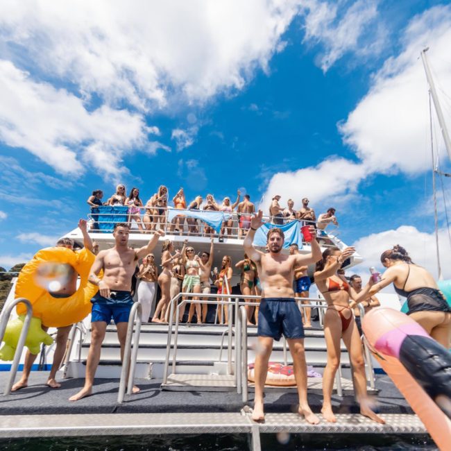 A group of people in swimwear stand and sit on a multi-deck boat under a bright blue sky. There are various colorful inflatable items around them, and another boat is visible in the background. Enjoy the thrill of a Sydney boat party hire on a private yacht charter Sydney Harbour.