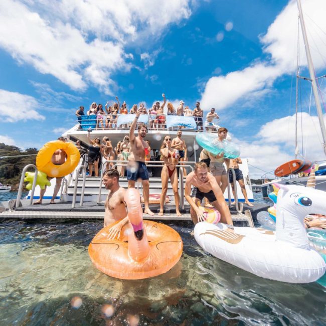A crowd of people on a catamaran party in Sydney; some are jumping into the water with inflatable floaties, while others watch from the deck.