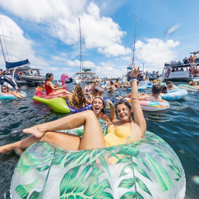 A group of people enjoying a sunny day on the water, floating on inflatable tubes and rafts, with boats anchored nearby. Two women in the foreground are smiling and waving. It's a perfect setting for a catamaran party Sydney style!