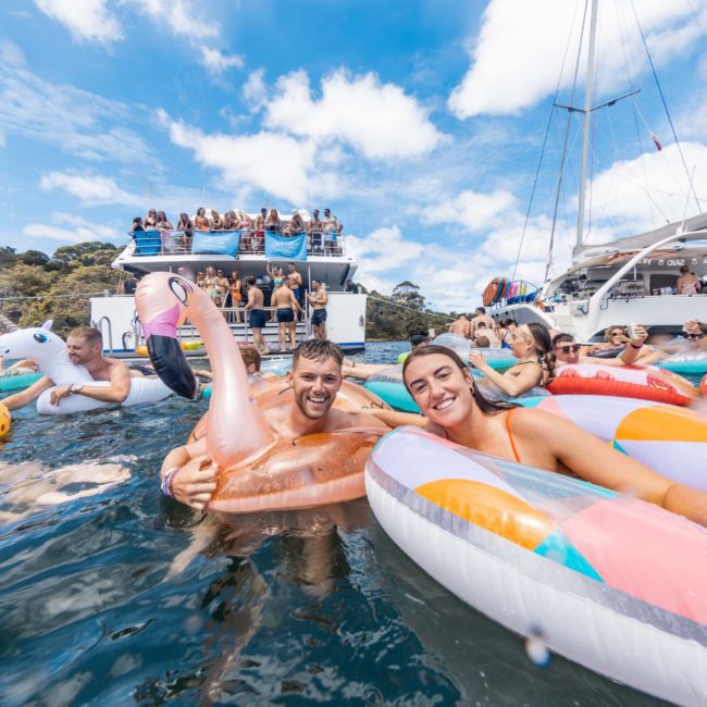 People enjoying a sunny day on the water, floating on inflatable pool toys near docked boats. There is also a crowded two-story boat visible in the background against a clear blue sky, offering a perfect setting for DJ boat hire Sydney or corporate boat events Sydney.