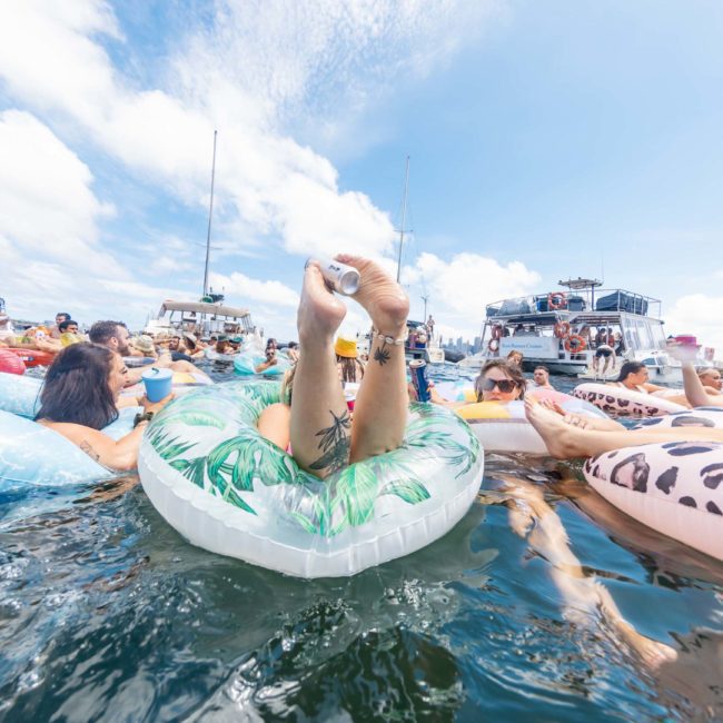 People floating in a lake on inflatable tubes, with boats and other people in the background. The sky is sunny with scattered clouds. For an elevated experience, consider a luxury yacht hire Sydney to enjoy the waters in style.