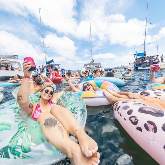 A group of people float on colorful inflatable tubes in the water, surrounded by boats under a blue sky with clouds. Some hold drinks, smiling and relaxing as they enjoy a Catamaran party in Sydney.