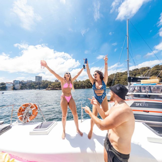 Two women in swimsuits stand on a private yacht charter on Sydney Harbour, raising their arms, while a man in a hat gestures towards them. Other boats and people are in the background on a sunny day near a wooded shore.