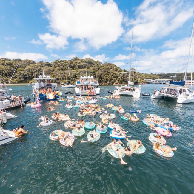 A large group of people in colorful inflatable rings float in the water surrounded by multiple boats on a sunny day with a forested shoreline in the background, enjoying a luxurious catamaran party Sydney.