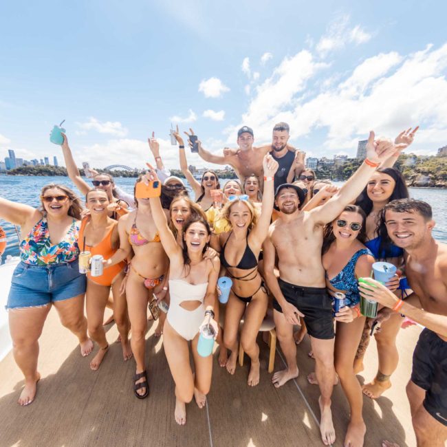 A group of people in swimsuits and casual attire are on a luxury yacht, smiling and raising their arms in celebration with the stunning Sydney skyline and water in the background.