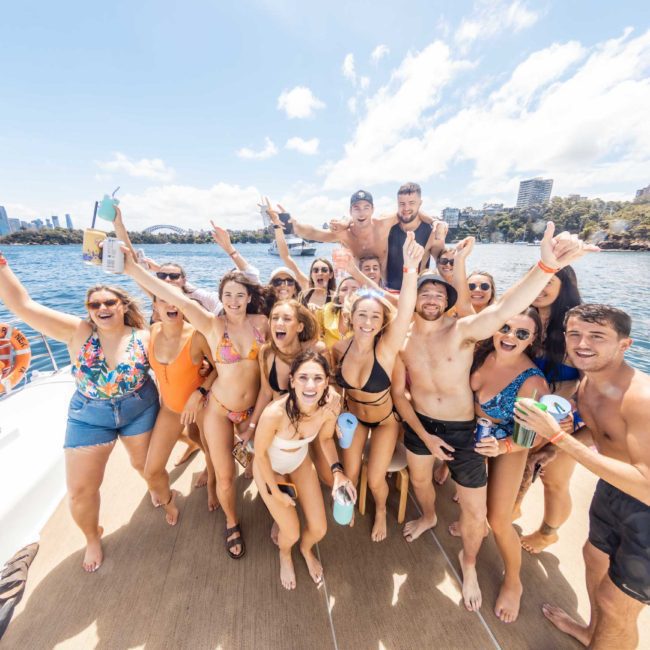 A group of people in swimwear pose energetically on a boat with a cityscape and waterfront in the background. They are smiling and raising their arms, appearing joyful and celebratory, enjoying what looks like an incredible private yacht charter on Sydney Harbour.