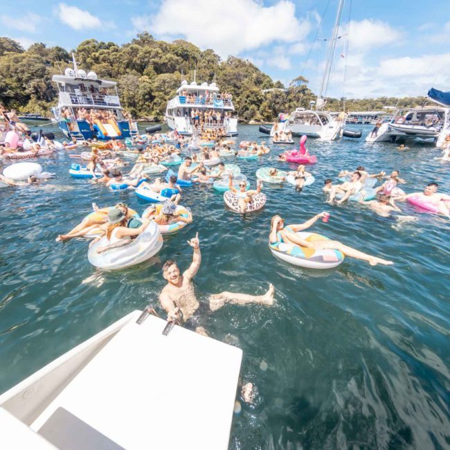 People are relaxing on inflatables in a crowded waterway surrounded by boats with trees in the background on a sunny day, enjoying what looks like a perfect Sydney boat party hire.