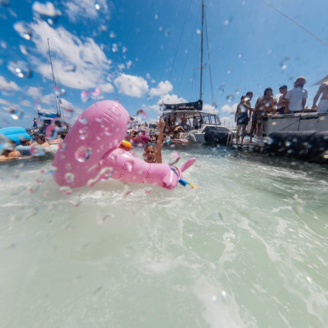 A person floats on a pink inflatable unicorn in the water, surrounded by people on boats and enjoying a Sydney boat party hire under a bright blue sky.