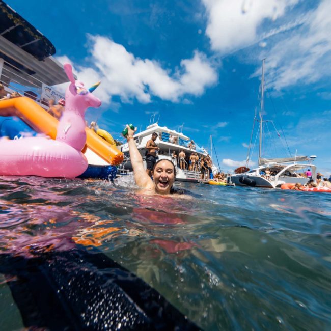 Person in the water smiling and raising a hand near a pink inflatable unicorn, surrounded by boats and people enjoying a sunny day on Sydney Harbour, ideal for a luxury yacht hire.