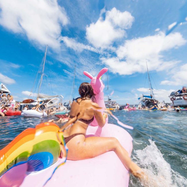 Person rides a colorful inflatable unicorn in a crowded, lively water area with boats and other people. It's sunny with a clear blue sky and some clouds. Nearby, there's an elegant luxury yacht hire in Sydney, adding to the vibrant scene.