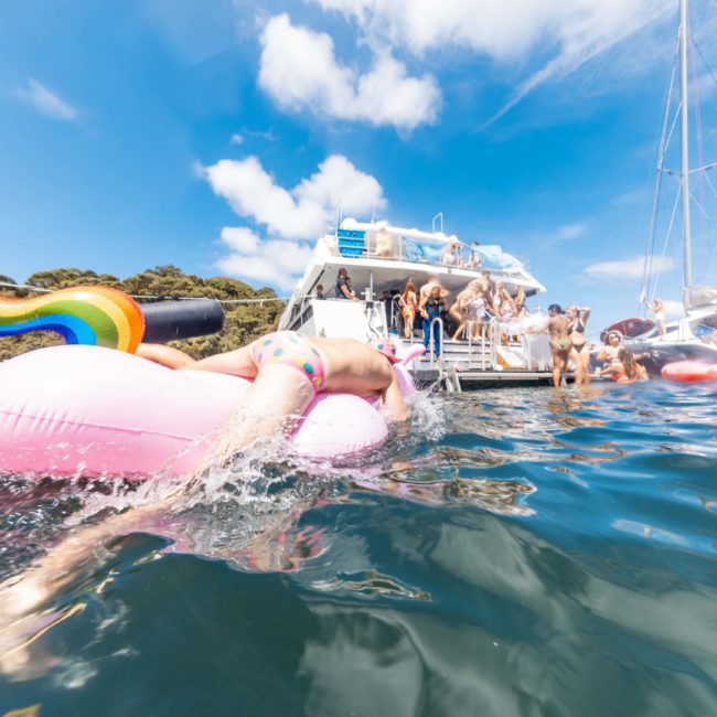 A person on a pink inflatable ring swims in the ocean near boats, under a clear sky. People enjoy themselves on luxury yachts and nearby vessels, making the most of Sydney's stunning waters.
