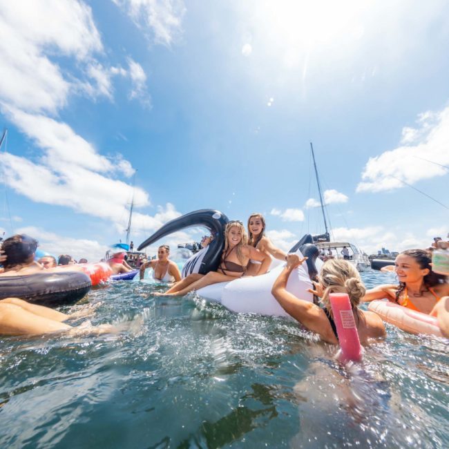 People on inflatable floats enjoy a sunny day in the water near boats with a bright blue sky above, creating a perfect ambiance for a Catamaran party in Sydney.