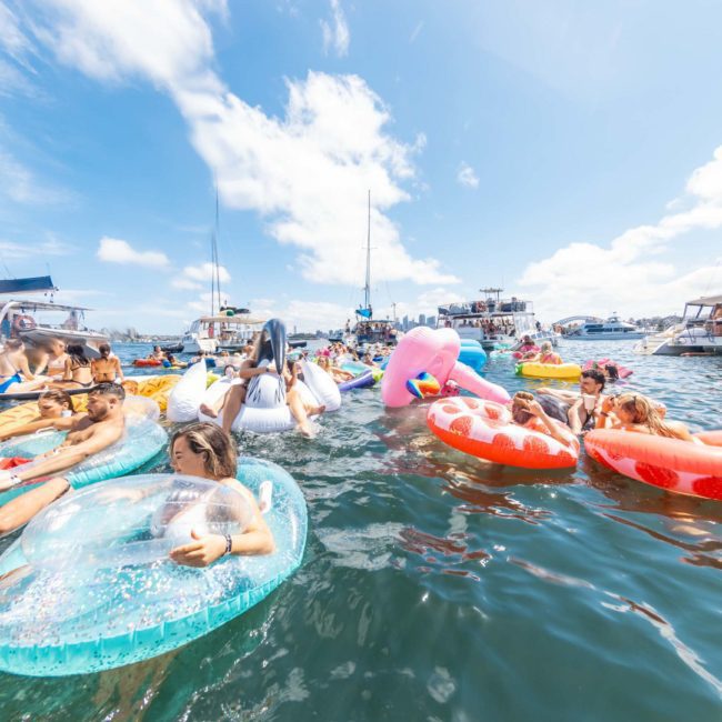 People on colorful inflatable floats gather in the water near several boats on a sunny day, enjoying a luxury yacht hire Sydney experience.