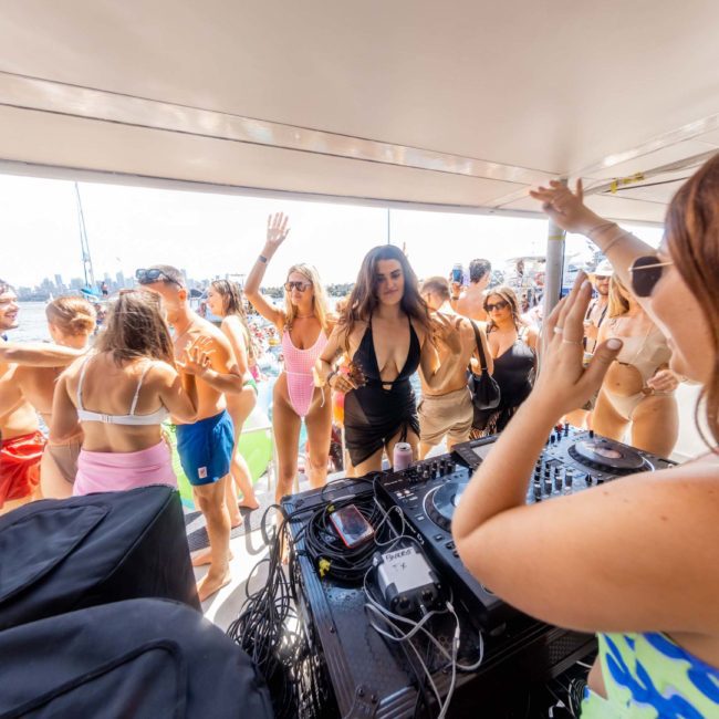 A group of people are dancing on a catamaran party in Sydney while a DJ plays music. Most individuals are wearing swimwear, and the scene is bright and lively.