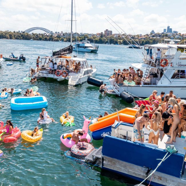People enjoying a sunny day on a lake with several boats and colorful inflatables, including a swan and flamingo. The background showcases a distant view of a bridge and city skyline, perfect for those considering luxury yacht hire Sydney or catamaran party Sydney adventures.