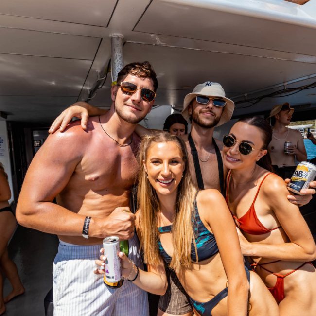 A group of young adults in swimwear enjoying drinks on a boat. Two men and two women are posing together, smiling at the camera, surrounded by others in the background. It looks like they’re having a fantastic time at a private yacht charter on Sydney Harbour.
