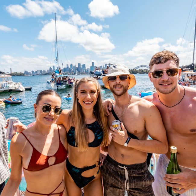 Four people in swimwear stand together and pose for a photo on a sunny day, with a city skyline and boats in the background. The scene hints at an unforgettable Sydney boat party hire experience.