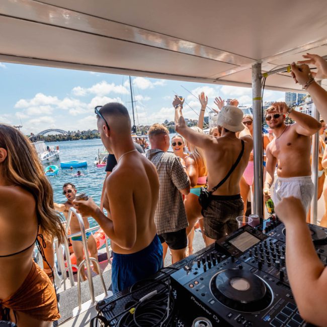 A group of people in swimwear dance and socialize on a luxury yacht hire in Sydney, featuring a DJ setup. The scene overlooks a body of water with buildings in the background under a partly cloudy sky.