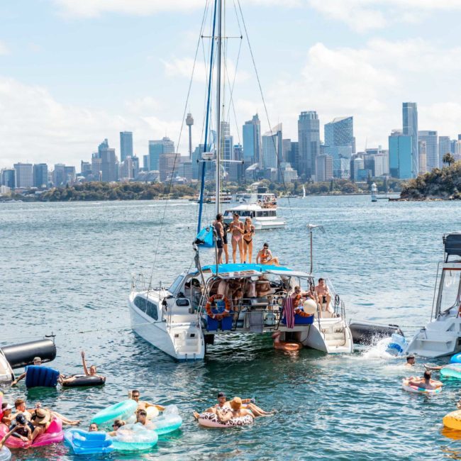 Boats and people on inflatable rafts in a harbor with a city skyline in the background, enjoying a catamaran party Sydney or corporate boat events Sydney.