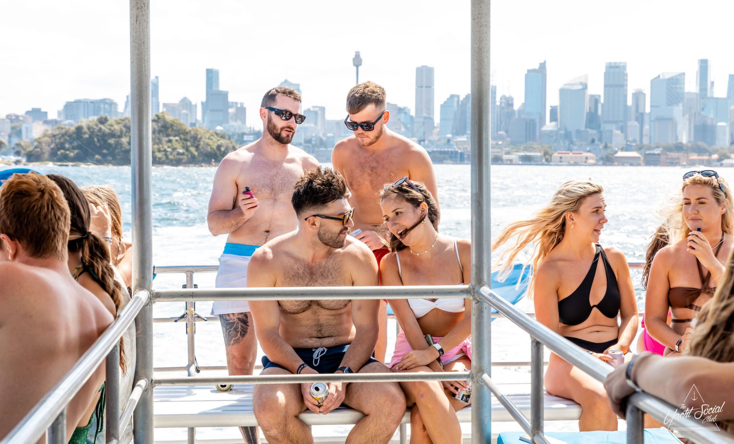 A group of people in swimwear are conversing and holding drinks on a luxury yacht in Sydney Harbour. A city skyline and water are visible in the background.