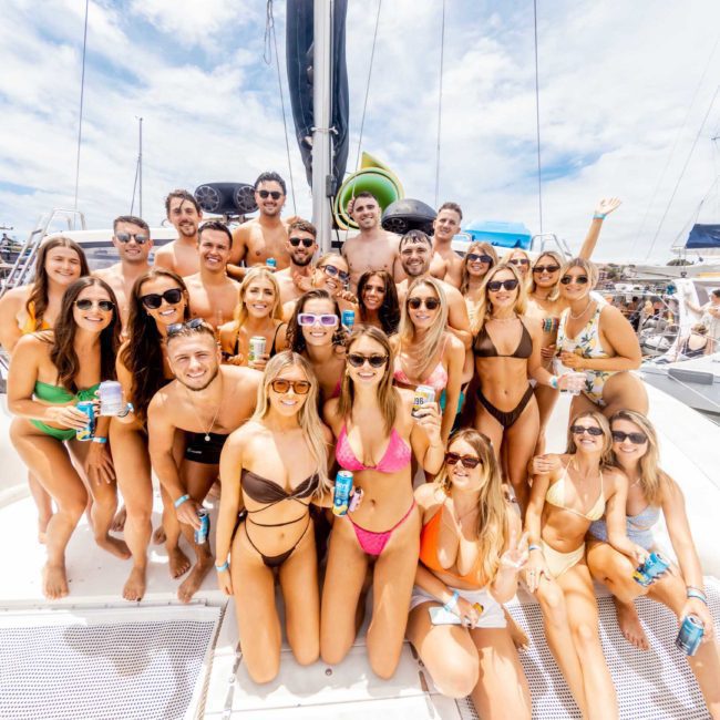 A large group of adults in swimwear poses for a photo on a catamaran party under a sunny sky in Sydney Harbour.