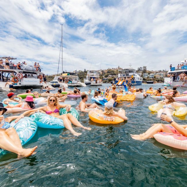 A group of people relax on inflatable floats in the water, surrounded by several anchored boats on a sunny day. It looks like the perfect setting for a Sydney boat party hire or even a private yacht charter on Sydney Harbour.