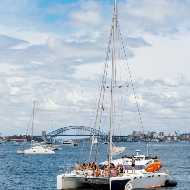A catamaran filled with people sails on a body of water with boats in the background and a bridge visible in the distance under a partly cloudy sky, epitomizing the allure of Luxury yacht hire Sydney.