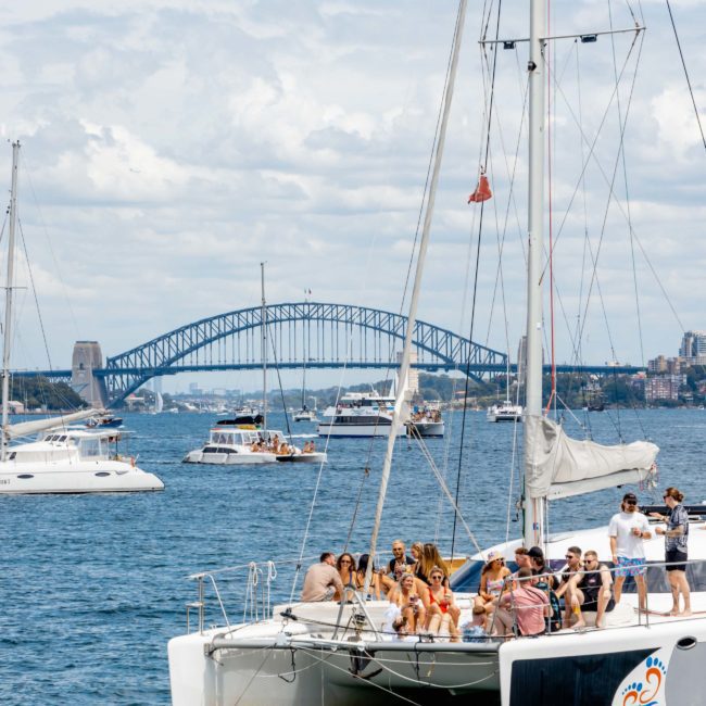 A group of people are sitting and standing on a luxury catamaran in a harbor with a large bridge and other boats in the background on a cloudy day.