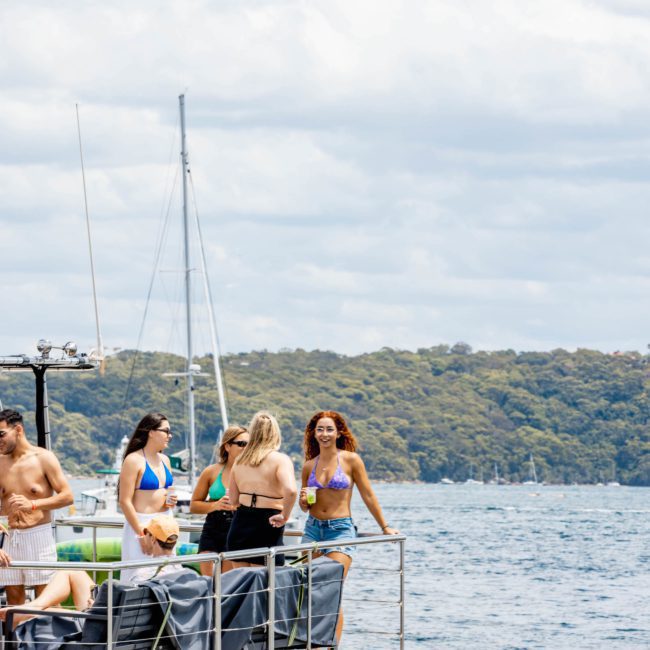 Group of people in swimwear standing and conversing on the deck of a boat on the water with trees and sailboats in the background, enjoying a DJ boat hire Sydney experience.