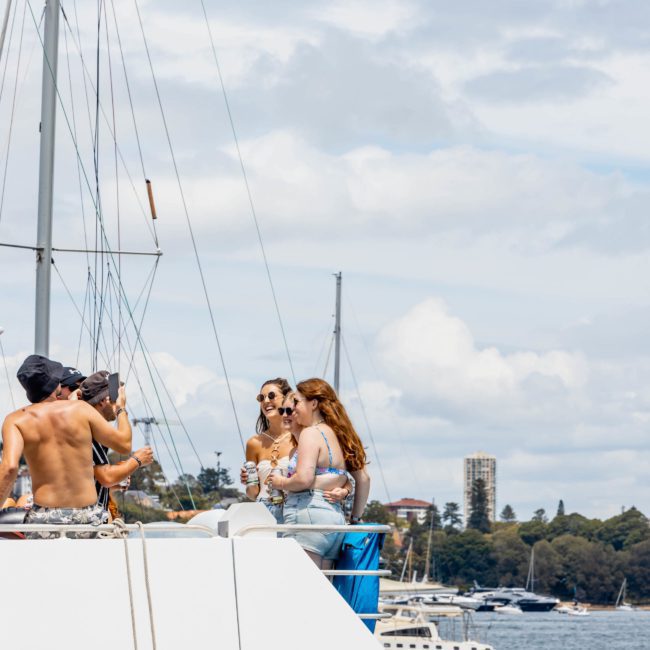A group of people on a boat, some standing and some seated with drinks in hand. Other boats and a distant shoreline are visible under a partly cloudy sky. Enjoy a memorable experience through a private yacht charter in Sydney Harbour or join the ultimate catamaran party in Sydney.