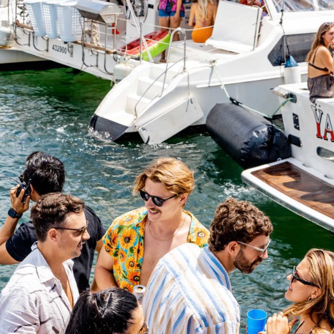A group of people socializing and drinking on a boat docked near the shore, with other boats and individuals in the background. Sunlight and bright attire suggest a casual, summer gathering. Consider enhancing your experience with a DJ boat hire in Sydney for an unforgettable party vibe.