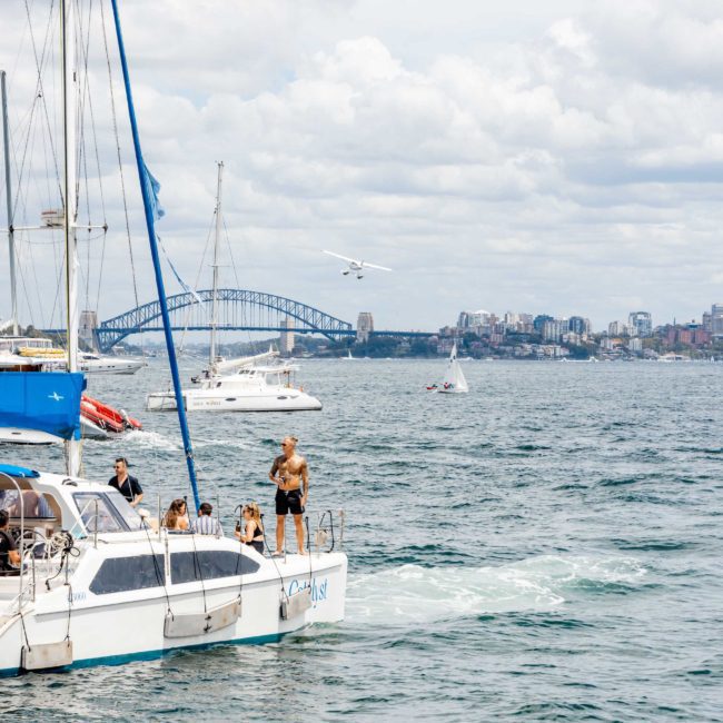 A group of people is on a catamaran party in Sydney near other boats and a visible bridge, with a seaplane in the sky. The sky is partly cloudy, and the scene appears to be set in a city with visible buildings.