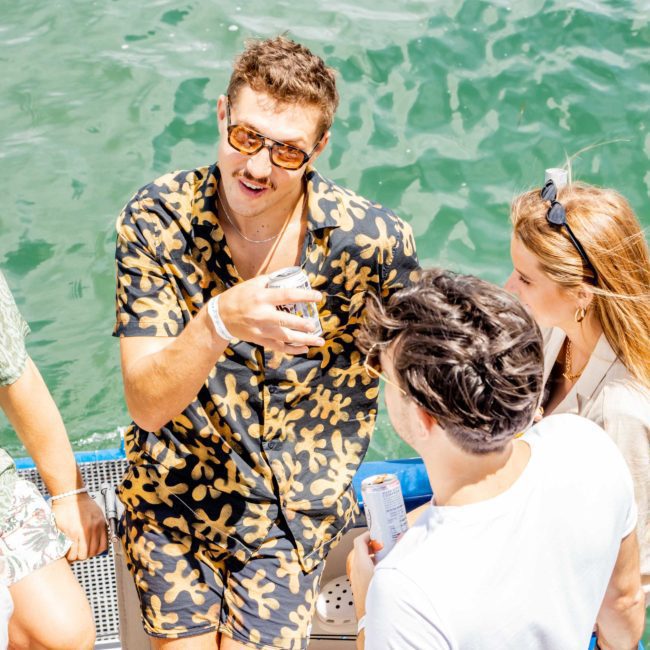 A person wearing a floral-patterned outfit holds a can while conversing with two others near a body of water during what seems like a lively Sydney boat party hire.