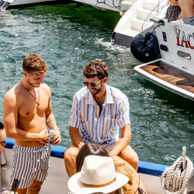 A group of people socializing on a boat. Two men in striped clothing are in the center, with one standing and holding a can and the other sitting. Water and another boat are visible in the background, perfect for a Sydney boat party hire or a luxury yacht hire in Sydney Harbour.