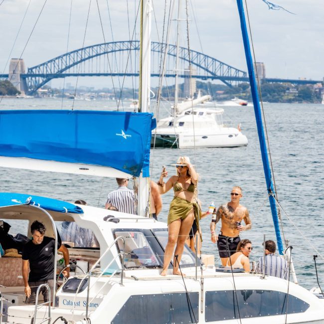 People on a catamaran enjoying a sunny day on the water with the Sydney Harbour Bridge visible in the background, taking in the perfect setting for a corporate boat event Sydney.
