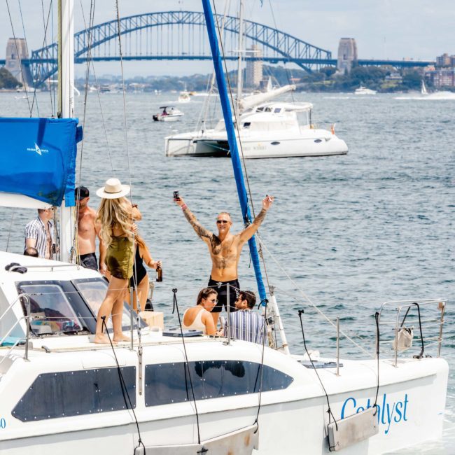 A group of people are on a sailboat named "Catalyst" in a body of water with other boats. A man stands with arms raised. A bridge is visible in the background under a cloudy sky, setting the perfect scene for a corporate boat event or catamaran party in Sydney.