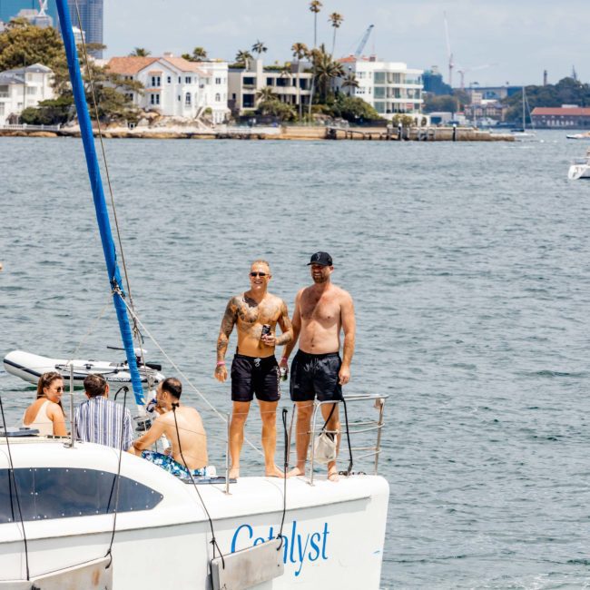 Two men stand on the bow of a sailboat named 'Catalyst,' with buildings and trees visible in the background on the shore, perfect for a Sydney boat party hire.