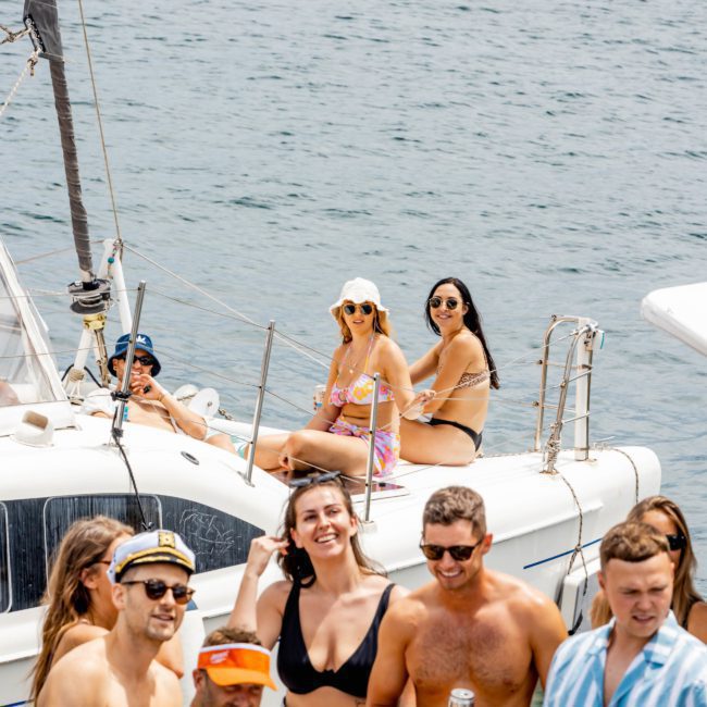 A group of people gathered on and around a boat on the water, some seated and some standing, enjoying a sunny day. Most are dressed in swimwear and holding drinks, relishing what seems to be a vibrant catamaran party Sydney style.