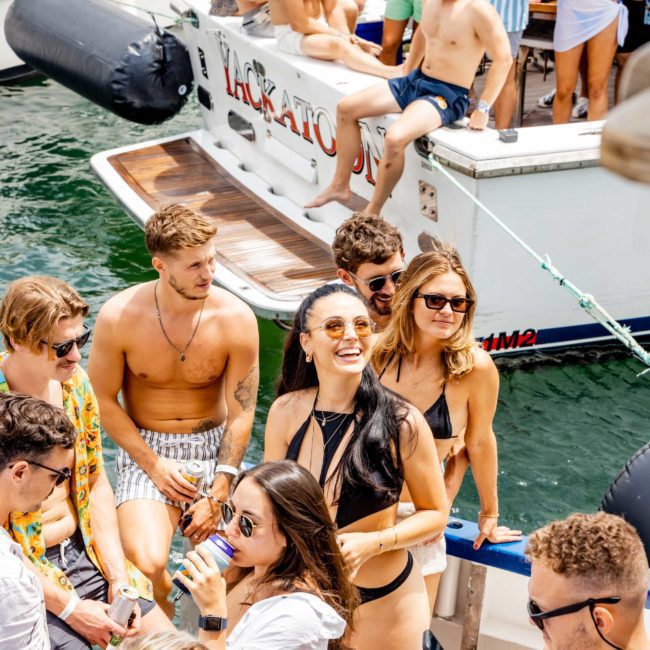 A group of people in swimwear socialize and enjoy drinks on luxury yachts docked together on a sunny day.