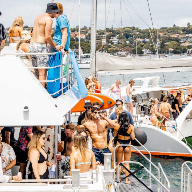 A group of people, dressed in swimwear, socializes on a docked boat. Some are standing on the upper deck, others on the lower deck, and more are on a nearby boat. The background shows water and a coastal area, providing the perfect setting for a luxury yacht hire Sydney experience.