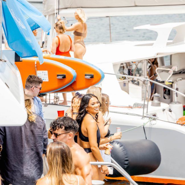 Several people are on a docked boat in sunny weather. Some are holding drinks, and others appear to be conversing. Two paddleboards are visible in the background, making it a perfect setting for Corporate boat events Sydney.