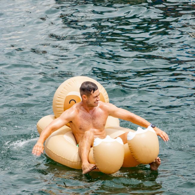 A man floats on a golden seashell-shaped pool float in a body of water while another person swims nearby, enjoying the luxurious atmosphere reminiscent of a private yacht charter in Sydney Harbour.