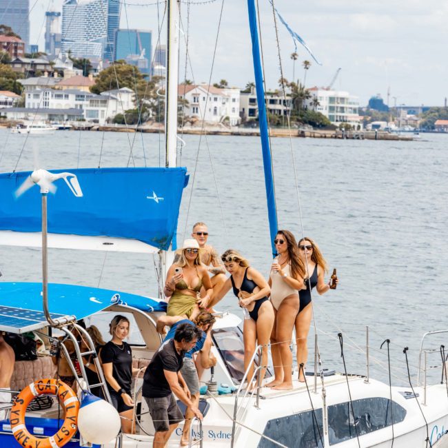 A group of people in swimsuits are on a private yacht charter Sydney Harbour and a catamaran, docked near a shore with buildings and cranes visible in the background.