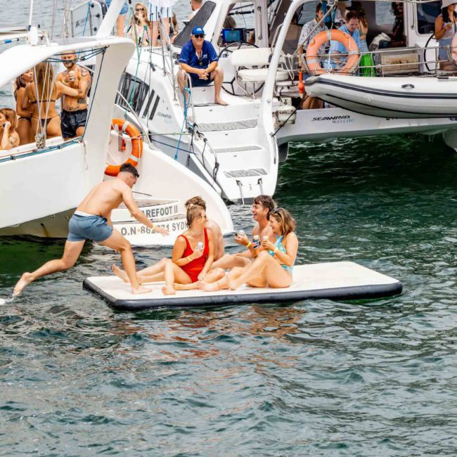 People are gathered on boats and a floating platform in a water setting. One person is jumping from a boat to the platform, while others are sitting and casually talking. It’s an ideal day for enjoying a private yacht charter in Sydney Harbour with friends.