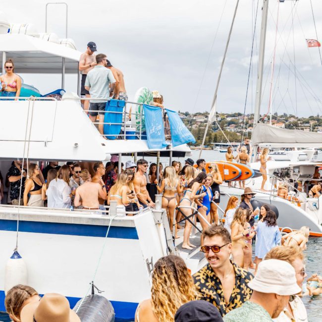 A vibrant catamaran party in Sydney with numerous people in swimwear standing, talking, and dancing on and around a large boat. Sailboats are visible in the background, adding to the festive atmosphere.