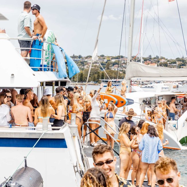 A large group of people is partying on a Sydney boat party hire and surrounding boats on the water. Many are wearing swimwear. The weather is cloudy yet bright, and the water is filled with boats creating a festive atmosphere.