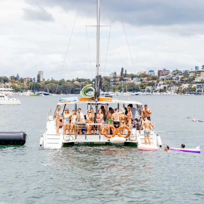 A group of people are gathered on a large catamaran in a harbor, enjoying a sunny day. Some are swimming nearby, with other boats and buildings in the background. It’s a perfect scene for corporate boat events or a Catamaran party in Sydney.